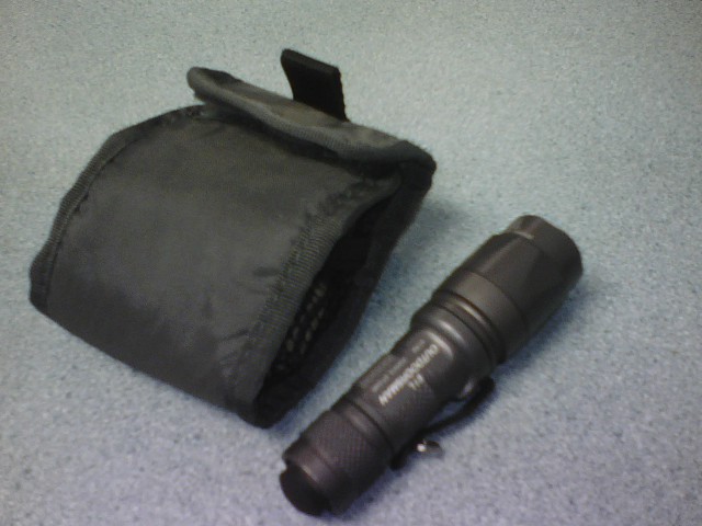 Black tactical flashlight with a black fabric case. Text on flashlight reads 'OUTDOORSMAN T18'