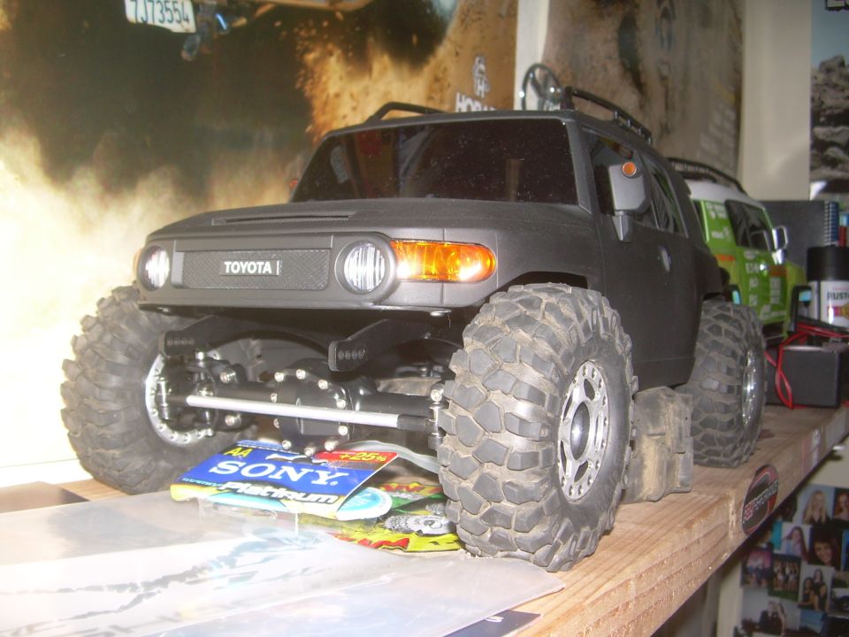 Black Toyota FJ Cruiser model with large off-road tires, front view. Another green vehicle is partially visible behind it