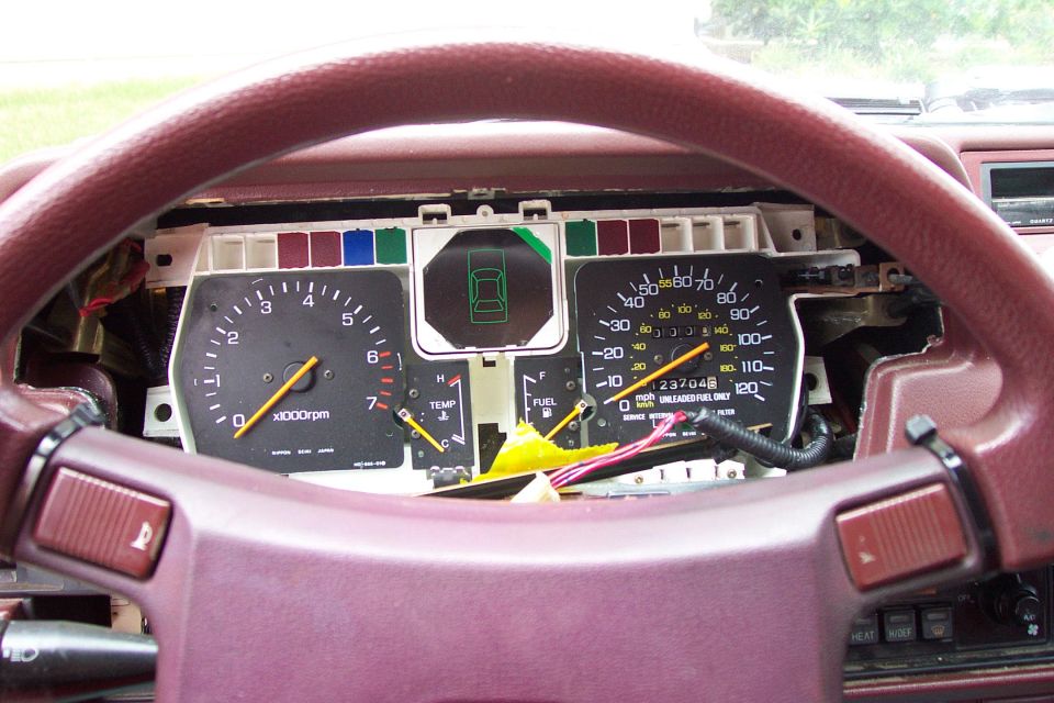Car instrument cluster with tach and speedometer viewed through a maroon steering wheel, wiring exposed