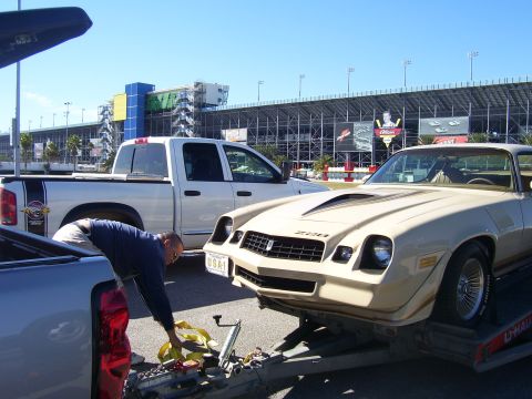 1979 Chevrolet Camaro - A man secures a tan Chevrolet Camaro Z28 onto a trailer, with a white pickup truck nearby and a racetrack grandstand in the background.