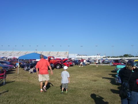 1979 Chevrolet Camaro - A wide view of a car show with many vehicles parked on the grass, people milling about, and a stadium in the background.