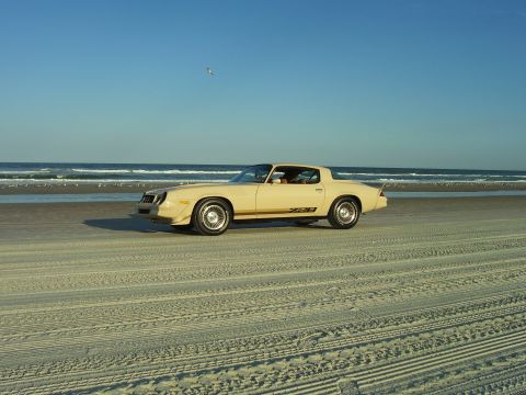 1979 Chevrolet Camaro - A tan 1979 Chevrolet Camaro Z28 is parked on the beach near the ocean under a clear blue sky.