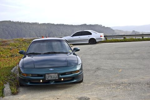 1996 Ford Probe - Two cars are parked on the side of a road with a scenic view of hills in the background.