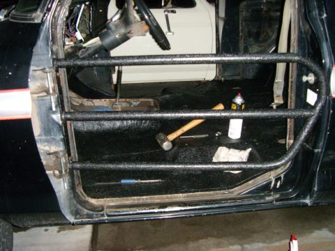 1978 Ford Bronco - A three-bar tube door finished in textured black bedliner hangs in the door opening of a gutted black SUV in a garage. Inside, the seats an…