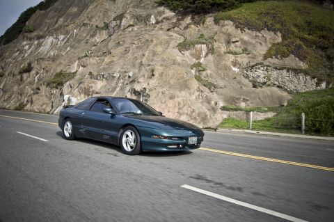 1996 Ford Probe - A teal 1996 Ford Probe GT drives along a scenic coastal highway with a large rock formation on the side.