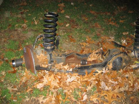 1996 Ford Bronco - Front suspension parts, including a coil spring and axle assembly, are scattered on the ground amidst fallen leaves.