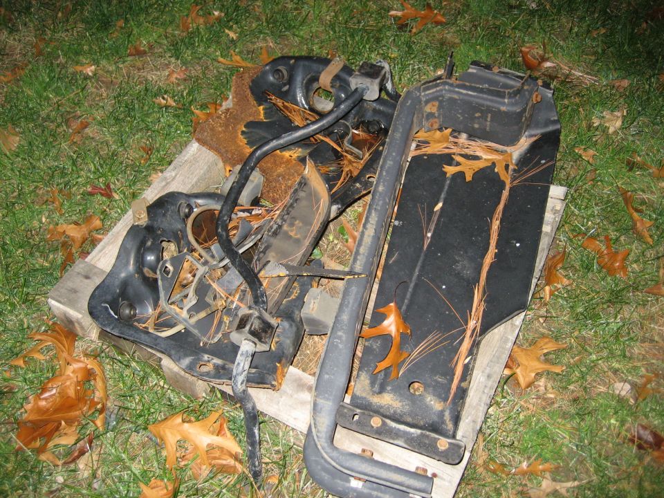 Rusted black metal vehicle components, possibly from an engine or suspension, lying on a wooden pallet outdoors with fallen leaves