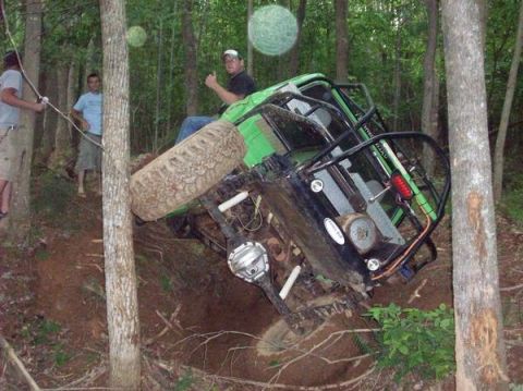 1978 Ford Bronco - A green off-road vehicle is tilted on its side in a wooded area, with a driver giving a thumbs up.