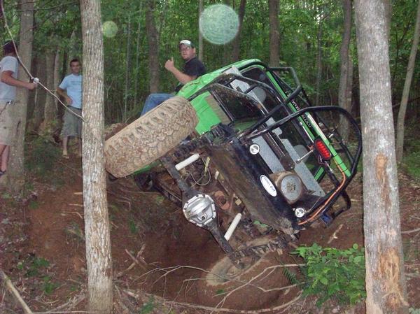 Green off-road vehicle on its side in woods, driver giving thumbs up