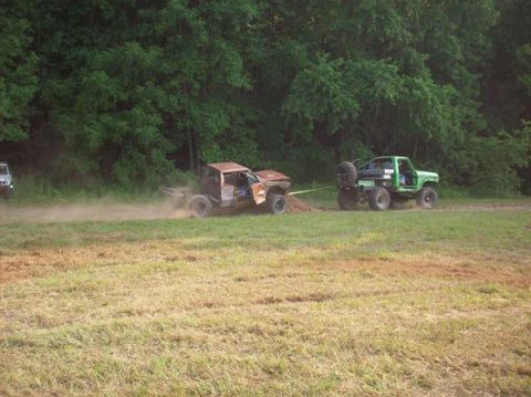 1978 Ford Bronco - A green off-road truck tows a damaged vehicle across a field with trees in the background.
