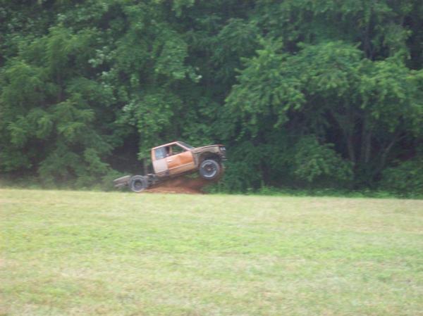 Pickup truck airborne over a dirt mound in a grassy field with trees in the background