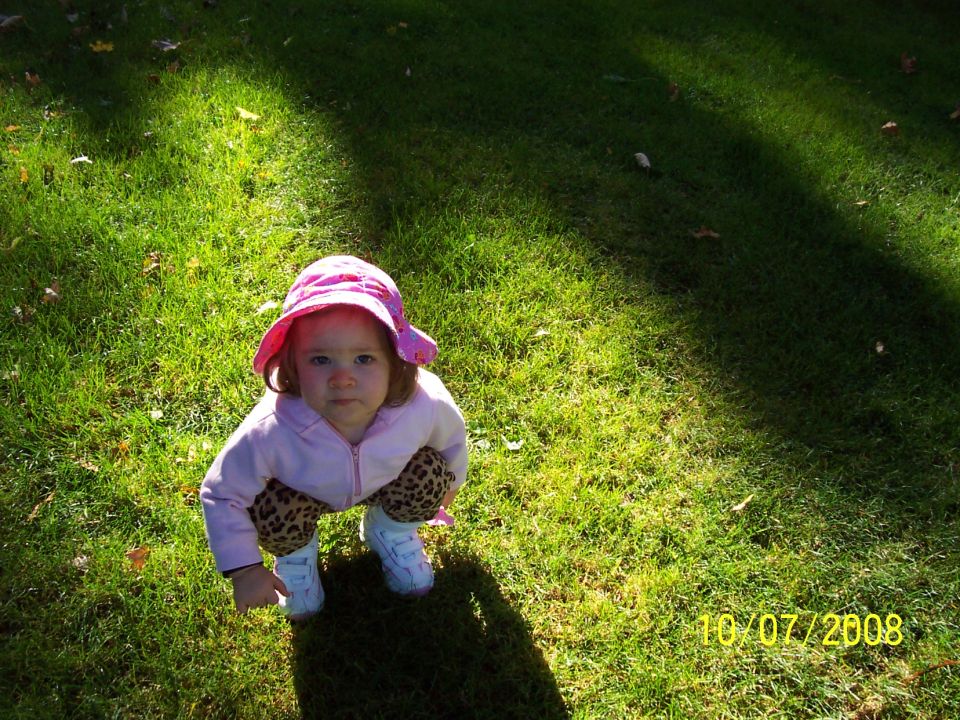 Toddler wearing a pink hat and leopard-print pants squats on grass. Date stamp reads 10/07/2008