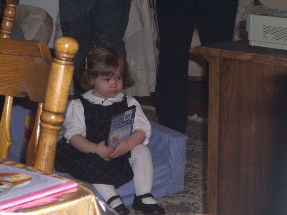 Toddler in a jumper dress and white tights sits holding a DVD case