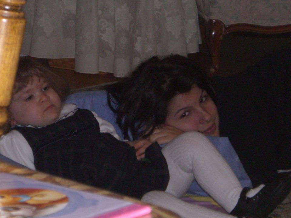 Child in dark overalls and white shirt lies next to a woman with dark hair, both on the floor