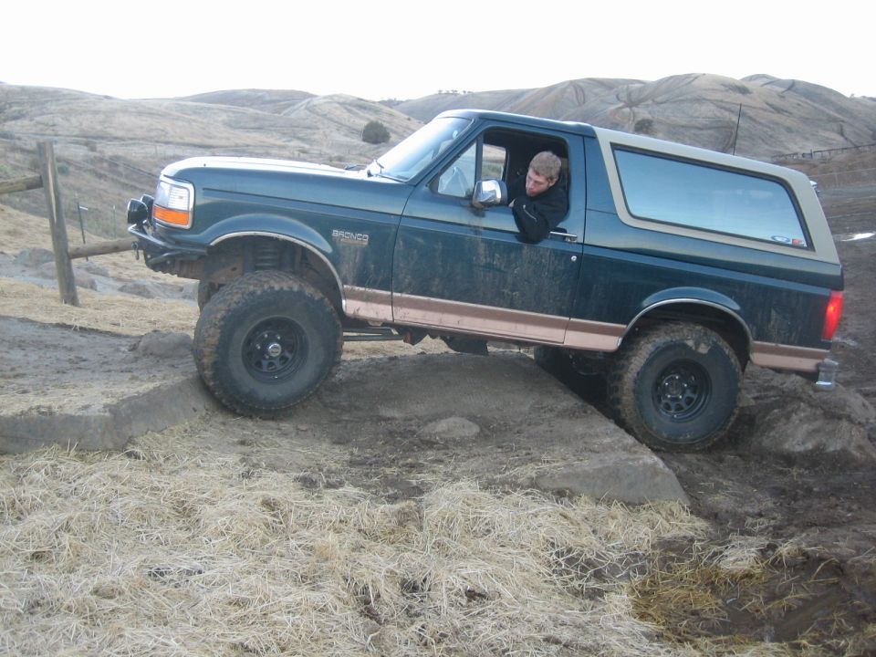Dark green 1995 Ford Bronco with tan top driving over rocks on a dirt trail