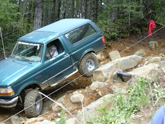 Green Ford Bronco with driver inside crawling over rocks between course ropes in the woods