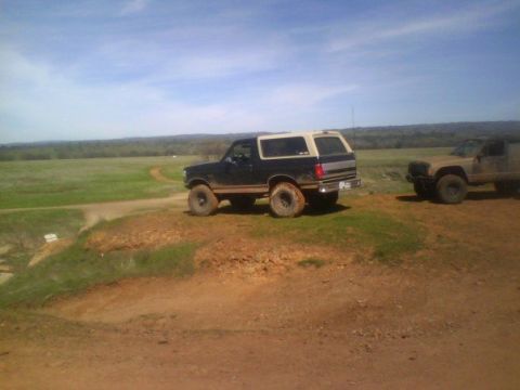 1995 Ford Bronco - A dark blue and tan Ford Bronco is parked on a dirt hill, with another SUV visible in the distance under a partly cloudy sky.