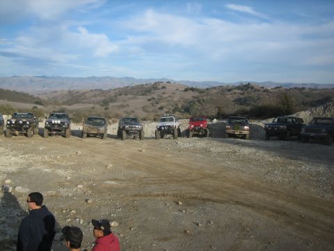 1995 Ford Bronco - A group of off-road vehicles are parked on a dusty lot with rolling hills under a blue sky.