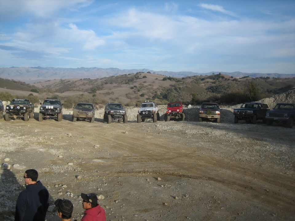 Several modified off-road trucks and SUVs parked on a dirt lot with hills in the background