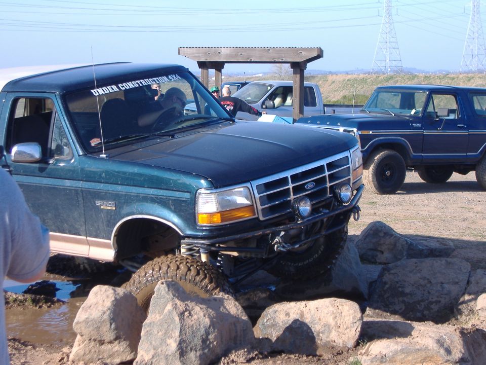 Green Ford Bronco navigating rocks, another Bronco and a pickup truck in background