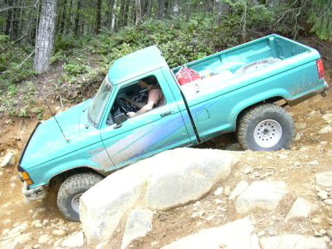 1991 Ford Bronco - A teal pickup truck is shown maneuvering over rocks on an off-road trail with trees in the background.