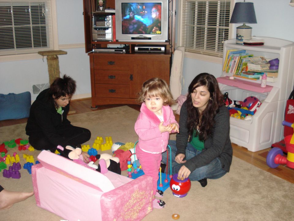 Two women and a toddler are in a living room with toys scattered around. A TV is on in the background