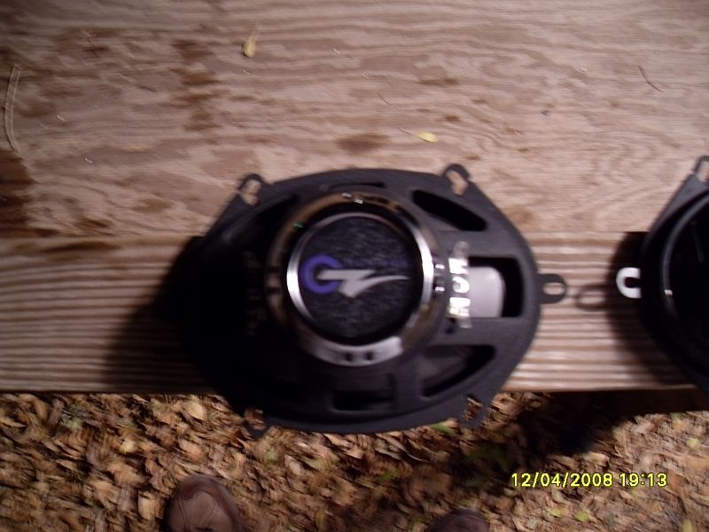 Two oval car speakers on a wooden surface, one with a silver and blue 'G' logo.