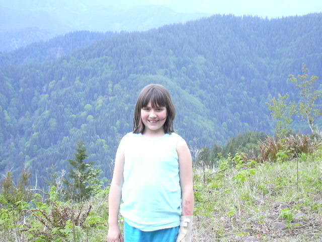Young girl in a light tank top smiling on a hilltop clearing with forested mountains behind