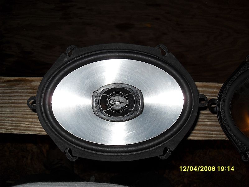 Two oval car speakers on a wooden surface. One speaker has a silver cone and black/silver logo.