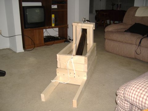 1988 Ford Bronco - A wooden contraption with a dark cylinder inside sits on a carpeted floor in front of a television stand and a plaid armchair.