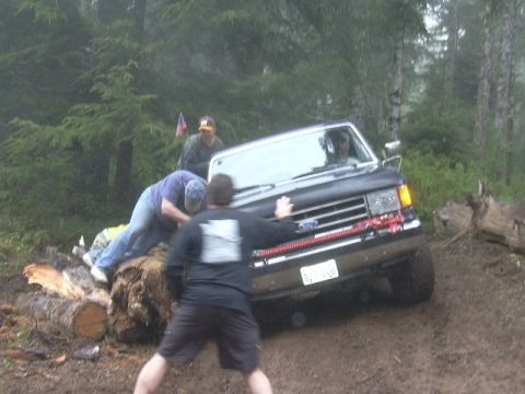 1991 Ford Bronco - People push a 1991 Ford Bronco stuck on logs in a forest setting.
