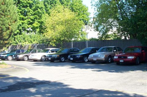 1991 Ford LTD Crown Victoria - A row of parked vehicles, mostly dark in color, sit under trees on a sunny day.