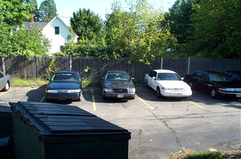 1991 Ford LTD Crown Victoria - Several Ford Crown Victorias are parked in a lot with dumpsters in the foreground and a house in the background.
