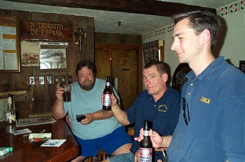 1991 Ford LTD Crown Victoria - Three men are gathered at a bar, holding beer bottles and glasses, with a sign reading 'UN TADASITO DE ESPANA' visible in the background.