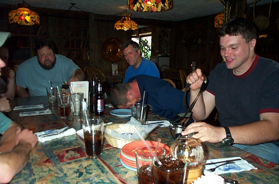 Group of men sitting at a table in a dimly lit restaurant, some with drinks, one with head down