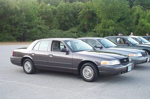 1991 Ford LTD Crown Victoria - A row of sedans are parked on asphalt, with trees in the background.