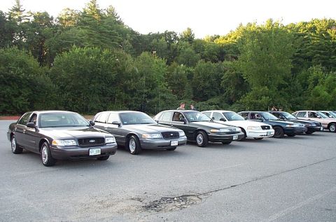 1991 Ford LTD Crown Victoria - Multiple Ford Crown Victoria police sedans are lined up in a parking lot with trees in the background.