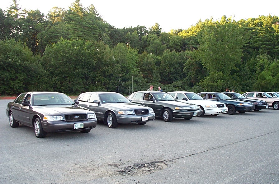 Several Ford Crown Victoria police cars parked in a line, facing right. The lead car is dark gray, followed by a lighter gray, then several white and dark sedans