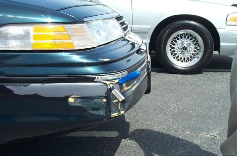 1991 Ford LTD Crown Victoria - A screwdriver is taped to the front bumper of a dark green sedan, with a silver sedan visible in the background.