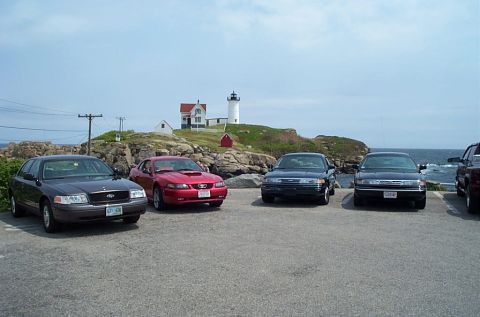 1991 Ford LTD Crown Victoria - Several cars are parked in a lot with a lighthouse and the ocean visible in the background.
