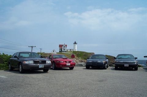 1991 Ford LTD Crown Victoria - Several cars are parked in a lot with a lighthouse and the ocean visible in the distance.