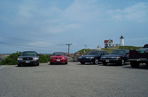 1991 Ford LTD Crown Victoria - A dark sedan, a red Ford Mustang, and two other vehicles are parked in a lot near a lighthouse.