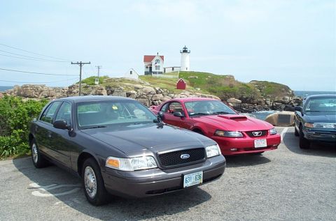 1991 Ford LTD Crown Victoria - Three cars are parked in a lot with a lighthouse and rocky coastline visible.