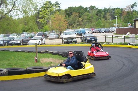 1991 Ford LTD Crown Victoria - Two bumper cars navigate a track at an amusement park, with a parking lot full of cars in the background.