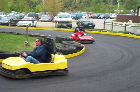 1991 Ford LTD Crown Victoria - Two go-karts navigate a turn on an outdoor track, with a parking lot visible behind the barrier.