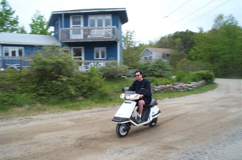 1991 Ford LTD Crown Victoria - A person rides a white scooter down a dirt road in front of a blue house.