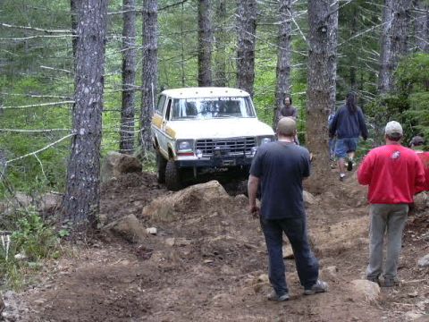 1991 Ford Bronco - An off-road vehicle is shown on a challenging, rocky path surrounded by trees, with several people observing.