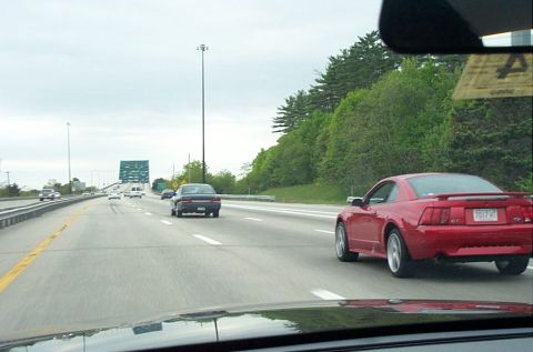 1991 Ford LTD Crown Victoria - A red Ford Mustang GT is seen driving on a highway with other vehicles under an overcast sky.