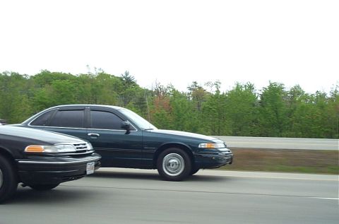 1991 Ford LTD Crown Victoria - Two dark-colored Ford Crown Victorias are seen driving on a highway with trees in the background.
