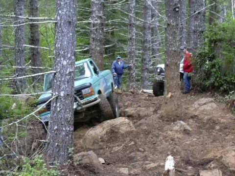 1991 Ford Bronco - Two people watch a teal Ford Bronco off-road vehicle climb over large rocks on a dirt path.